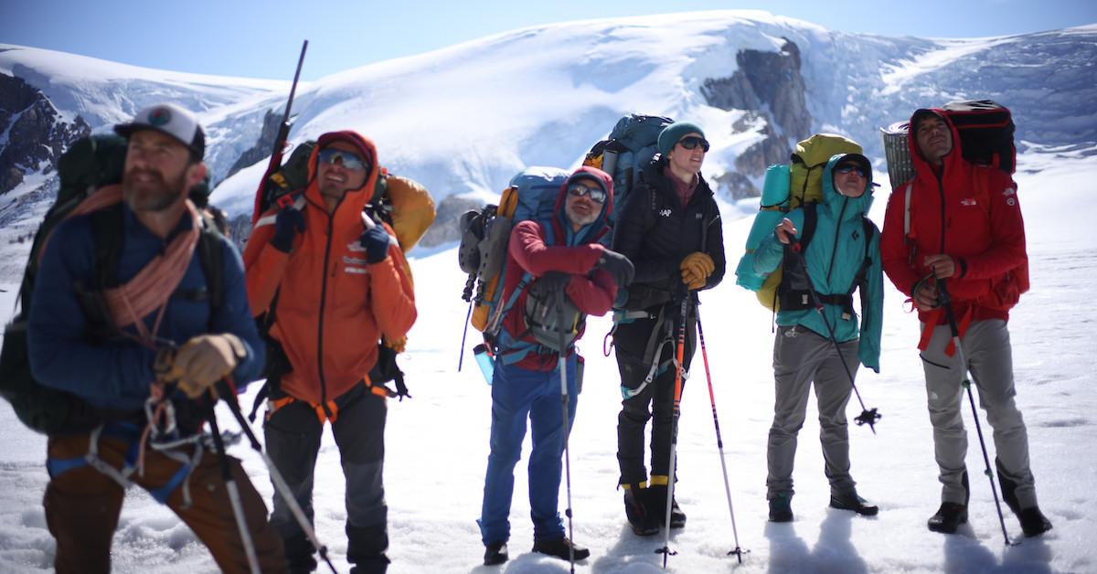 (Left to right): Aldo Kane, Adam Mike Jeldsen, Mikey Schaefer, Heidi Sevestre, Hazel Findley, and Alex Honnold wearing snow climbing gear pose by a snowy mountain.