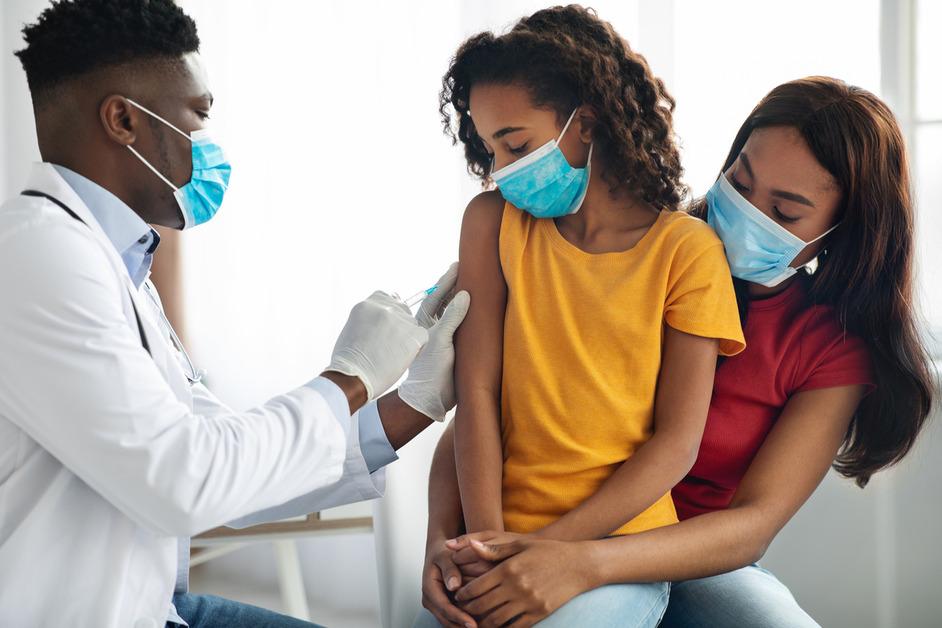 A daughter sits on her mothers lap while receiving a vaccine from a male doctor. 