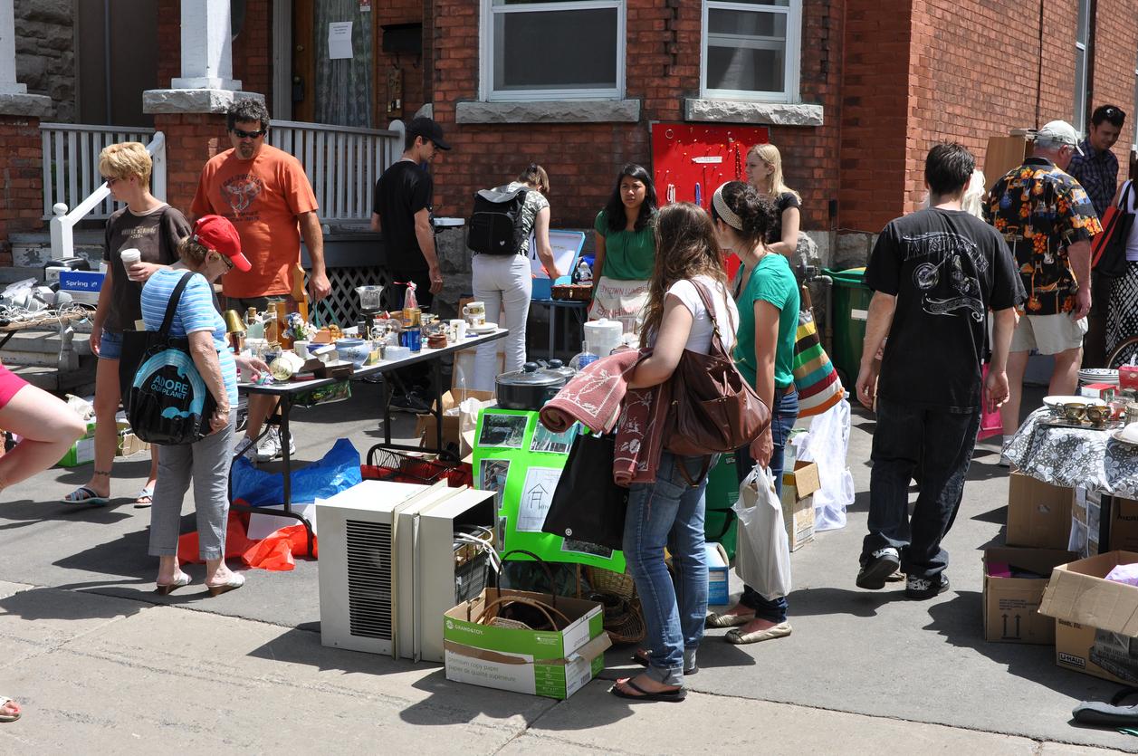 People carrying bags and shopping around tables of items at a garage sale.