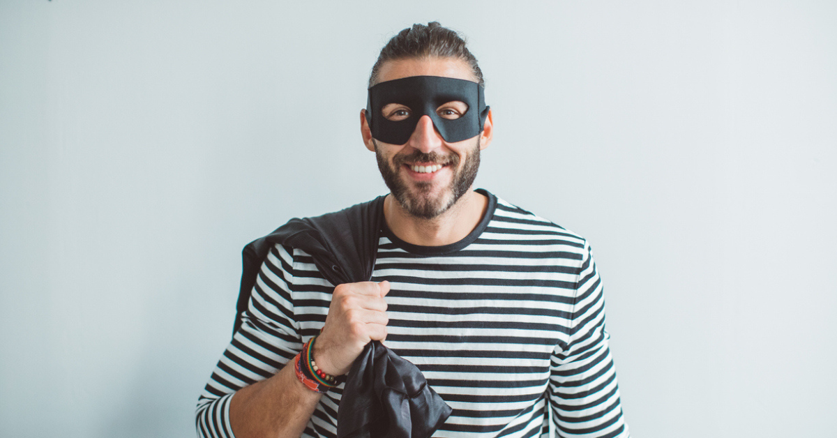 A man wearing a black bandit mask, black and white striped shirt, and carrying a black sack while dressed like a burglar 