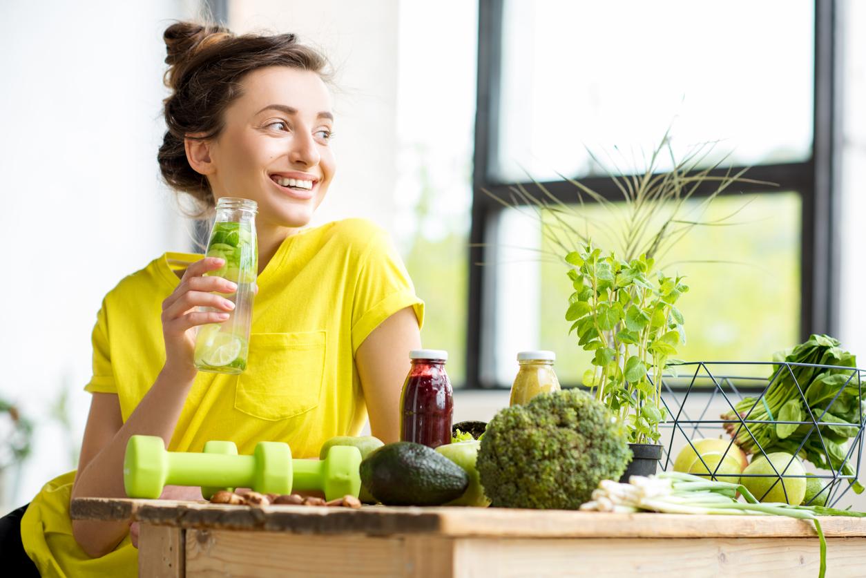 A smiling woman in a yellow top consumes a bottle of water infused with cucumbers with vegetables and dumbbells surrounding her.