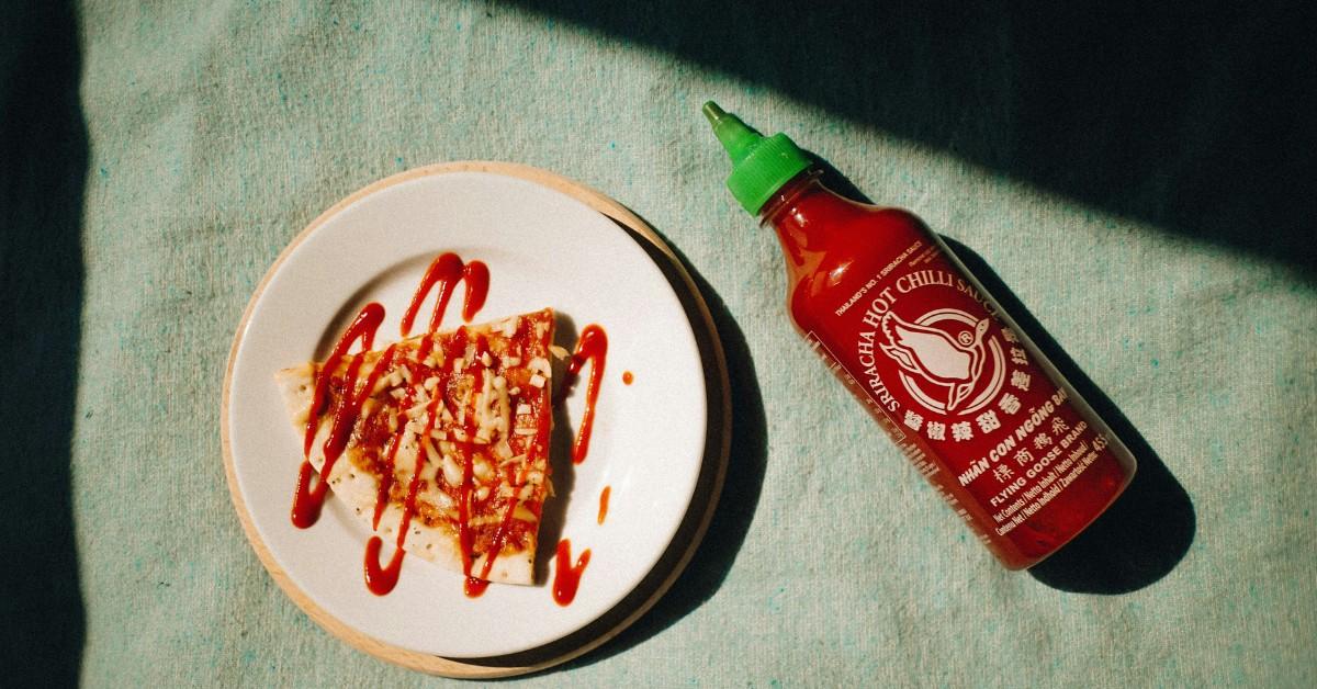 A bottle of Sriracha sits next to a slice of pizza