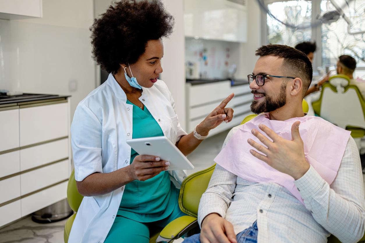 A dentist holding a tablet discusses the price of dental surgery with a patient laying in a chair with a pink bib.