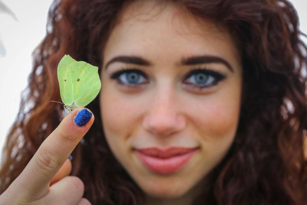 A woman with blue fingernail polish and blue eyes holds a green butterfly on the tip of her finger.