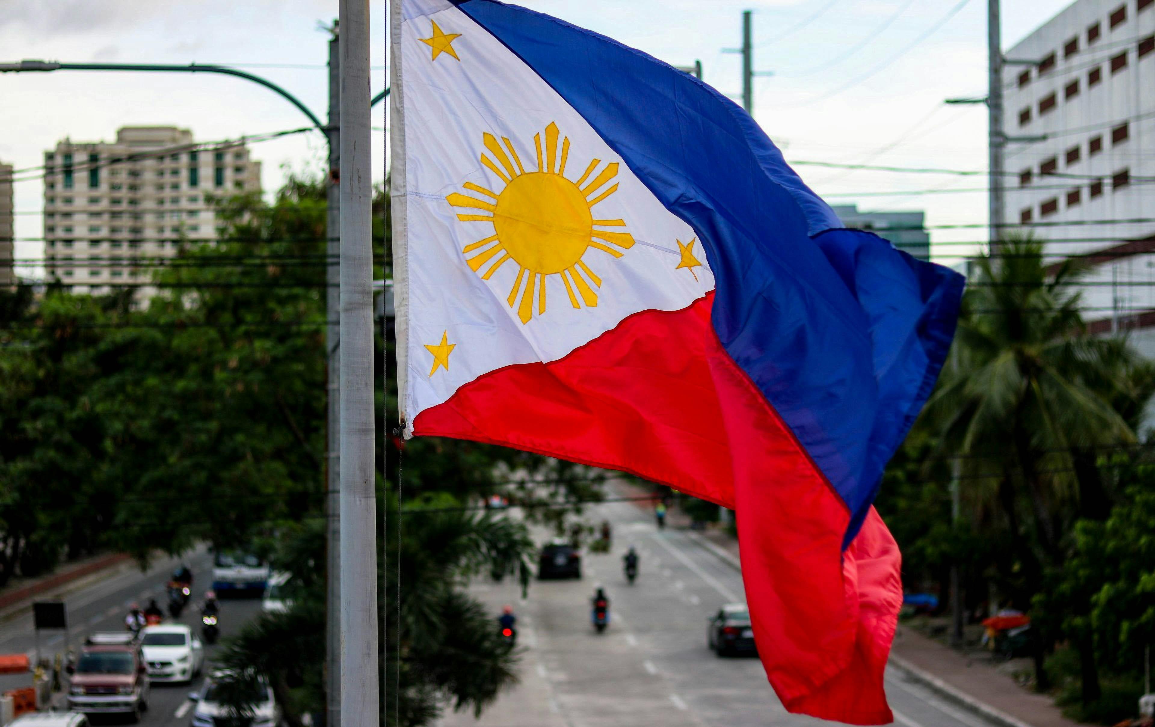 The flag of the Philippines is pictured in the center of a neighborhood with buildings on one side and a tree-lined street in the center.