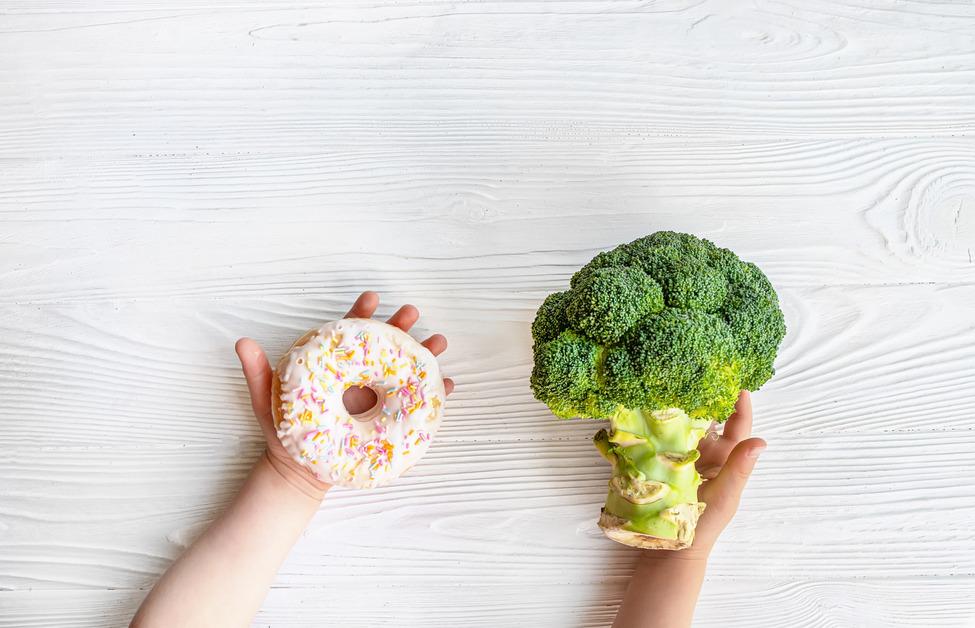 A person holding a white sprinkled donut next to a large bunch of broccoli in front of a white wood background. 