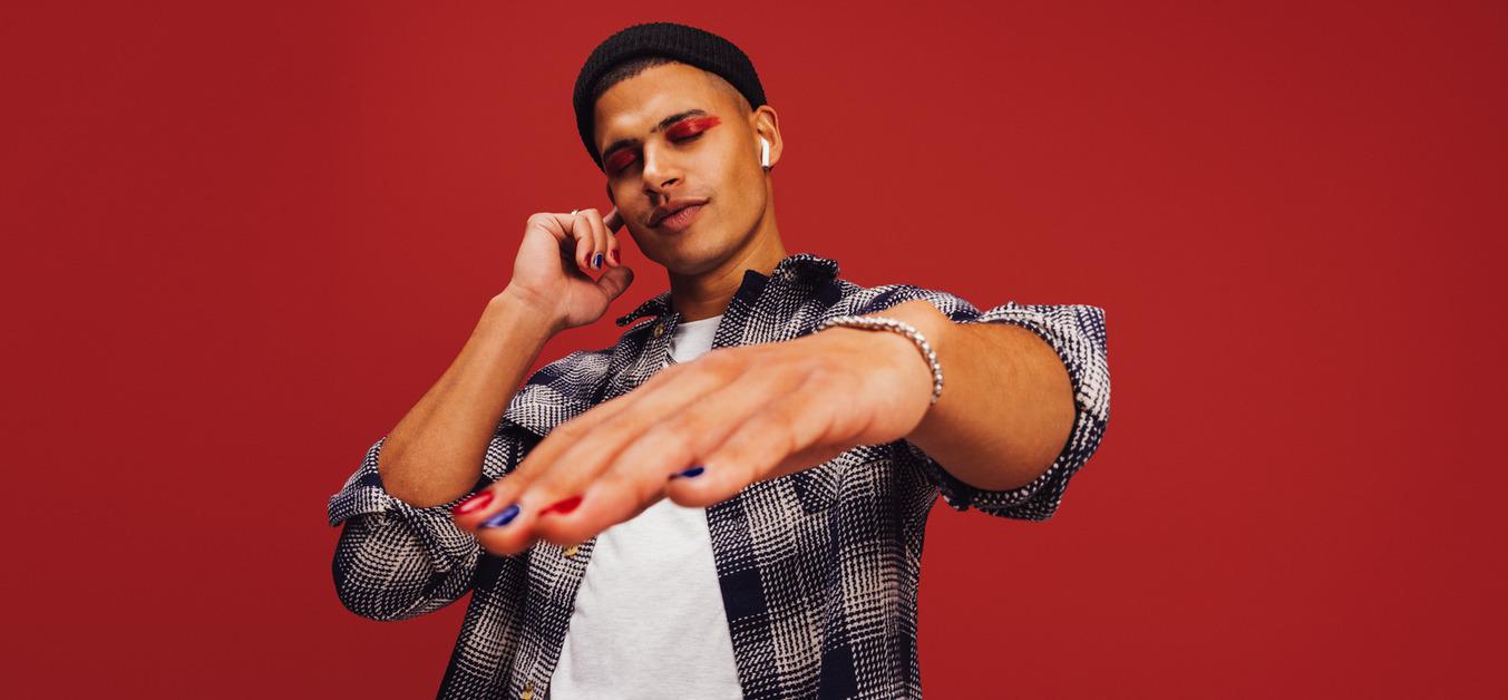 A man wearing a black beanie and flannel shirt stands in front of a red wall while holding one hand up to the AirPod in his ear and stretching out a hand near the camera with painted nails while closing his eyes.  