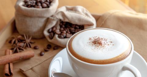 A cappuccino is placed on a plate beside coffee beans and cinnamon sticks.