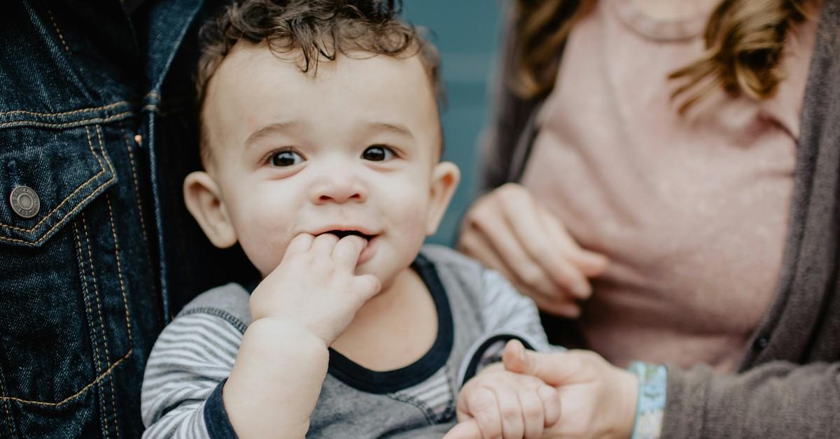 A toddler chews on his hands while sitting between his parents