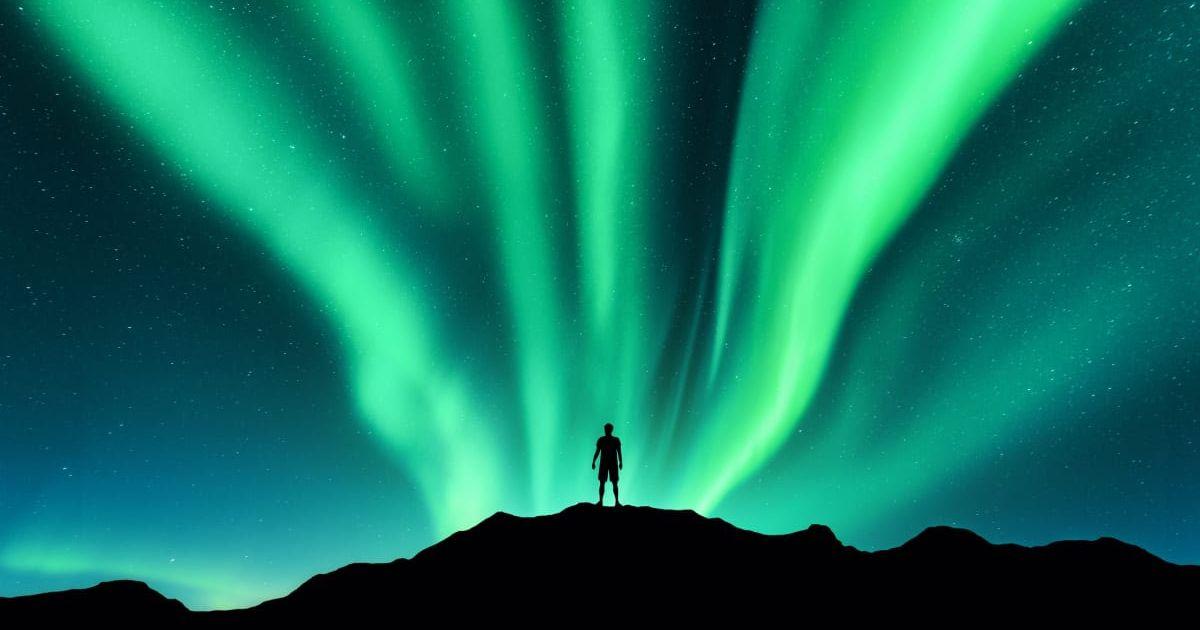 Silhouette of a man standing on a mountain beholding luminous green curtains of glowing lights decorating the night skies of Norway's Lofoten Islands during an aurora display (Representative Cover Image Source: Getty Images | Den Belitsk)