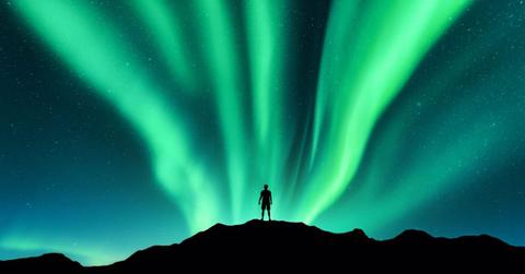 Silhouette of a man standing on a mountain beholding luminous green curtains of glowing lights decorating the night skies of Norway's Lofoten Islands during an aurora display (Representative Cover Image Source: Getty Images | Den Belitsk)