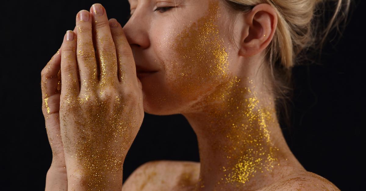 A closeup of a woman with gold glitter on her face, neck, and hands.