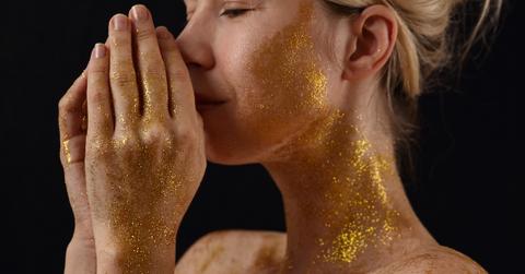 A closeup of a woman with gold glitter on her face, neck, and hands.