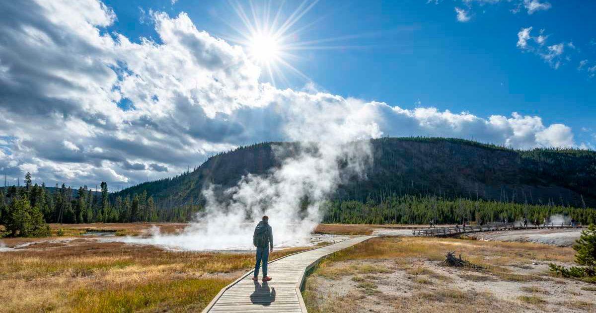 A tourist walks in front of a steaming hot spring in Yellowstone National Park (Representative Cover Image Source: Getty Images | imageBROKER | Mara Brandl)