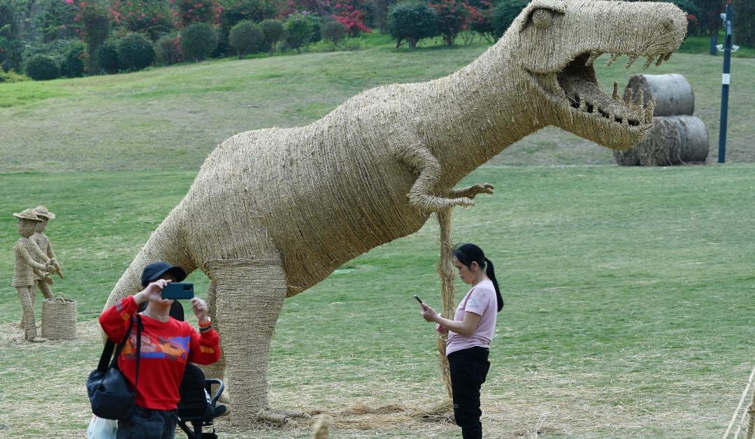 Two tourists are standing in front of a straw sculpture featuring a dinosaur during an art exhibition on March 6, 2023 in Nanning, Guangxi Zhuang Autonomous Region of China