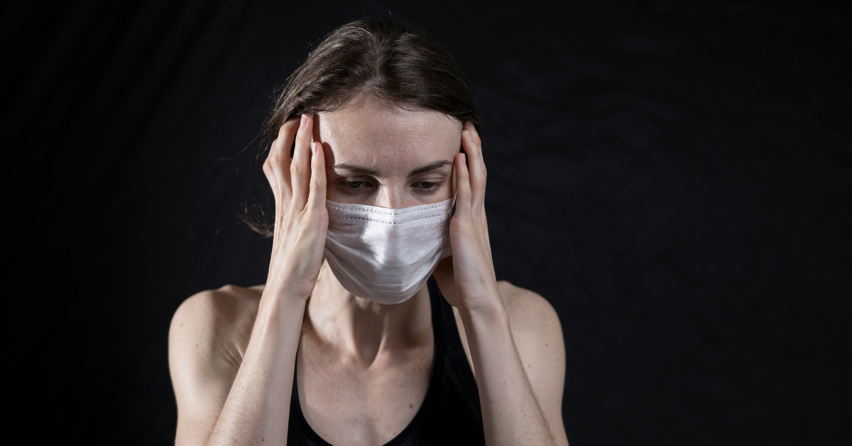 A woman holds her head while wearing a mask