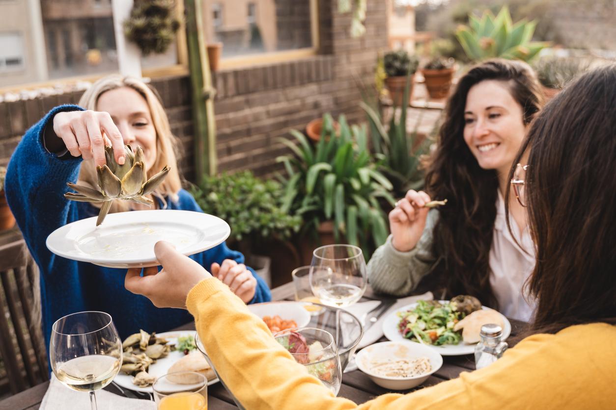 A group of three friends dine outdoors while one friend in a blue sweater holds an artichoke dangling above a plate.