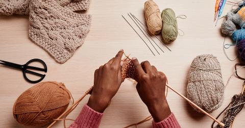 A person knitting at a table with skeins of yarn and knitting supples around the.