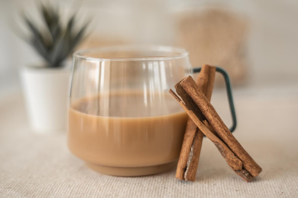 A cup of coffee is placed beside cinnamon sticks with a plant in the background.