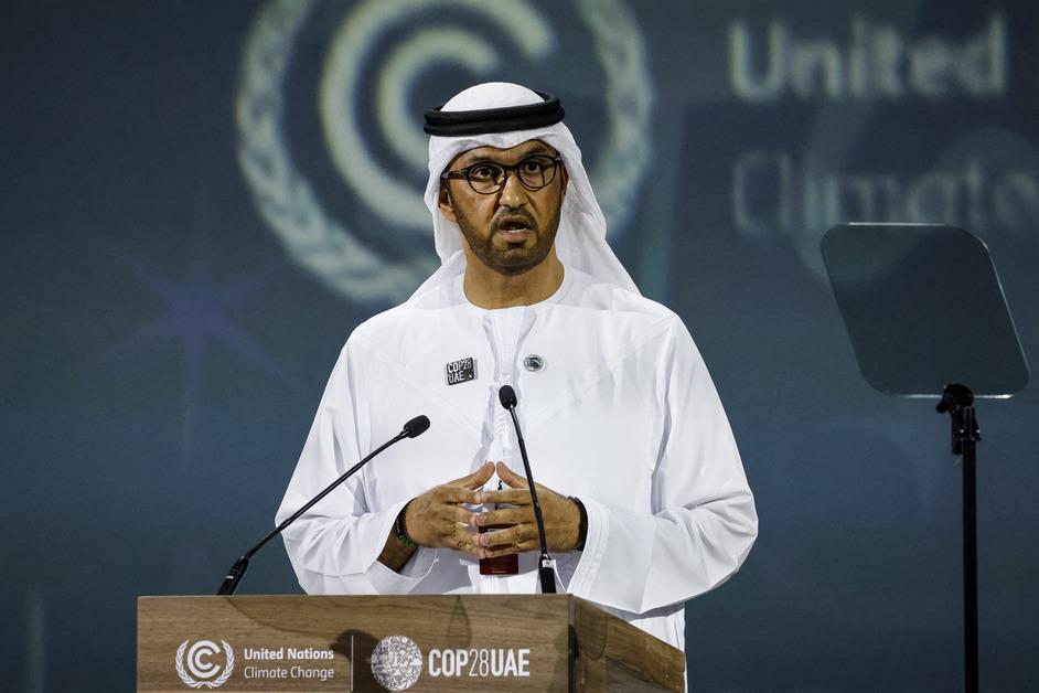 COP28 president Al Jaber stands at a podium at COP28 with his hands together while speaking and wearing a pin that says "COP28 UAE"