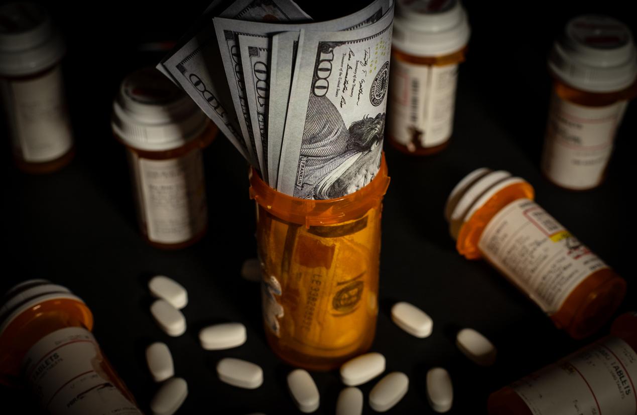 $100 bills are stuffed into an orange prescription drug container with white pills and other prescription bottles surrounding it.