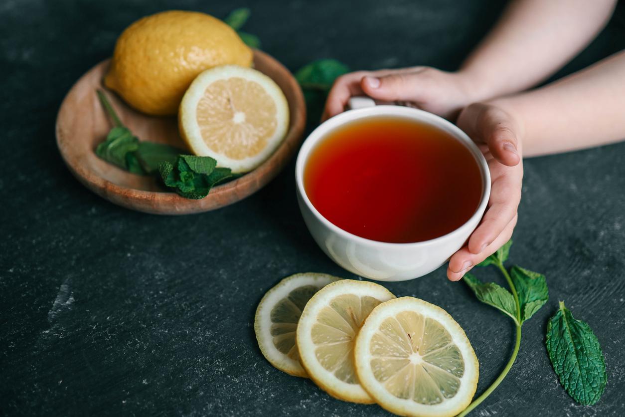 A person holds a large white cup of tea beside mint leaves and full and cut lemons.