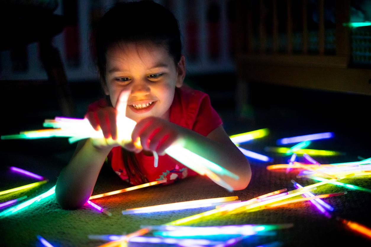 A young smiling child holds several glow sticks in their hands while glow sticks of various colors are placed around the child on the floor.