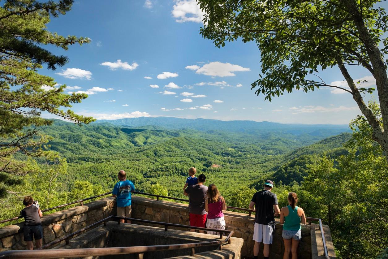 Tourists at a scenic outlook in the Smoky Mountains that overlooks green foliage, mountains, and blue skies.