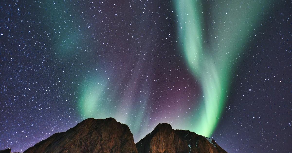The greens and pinks of the northern lights swirl above a mountain