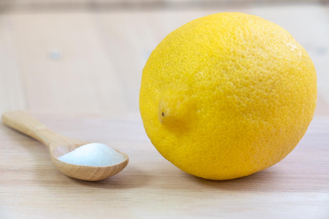 A close-up photo of a wooden spoon with salt granules beside a whole lemon atop a wooden cutting board.