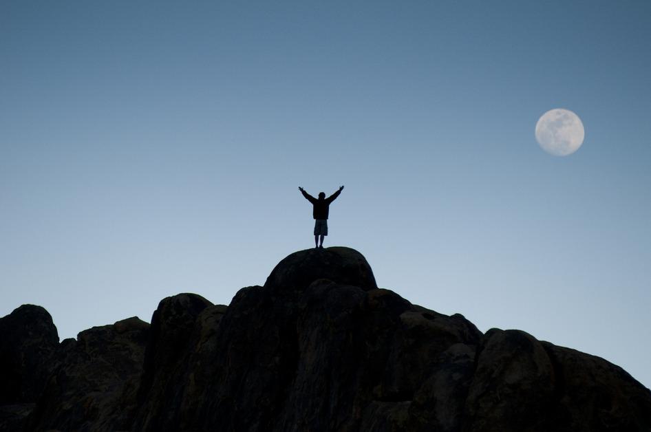 A person stands on a rock facing the sky with a full moon. 