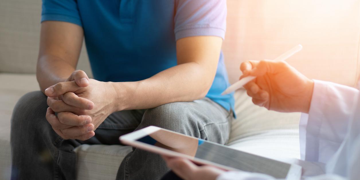 A low-angle view of the body of a man sitting on a couch clasping his hands while a doctor takes notes on an iPad.