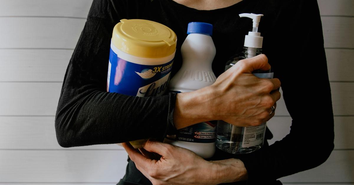 Woman in black shirt holding cleaning wipes, bleach, and hand sanitizer.