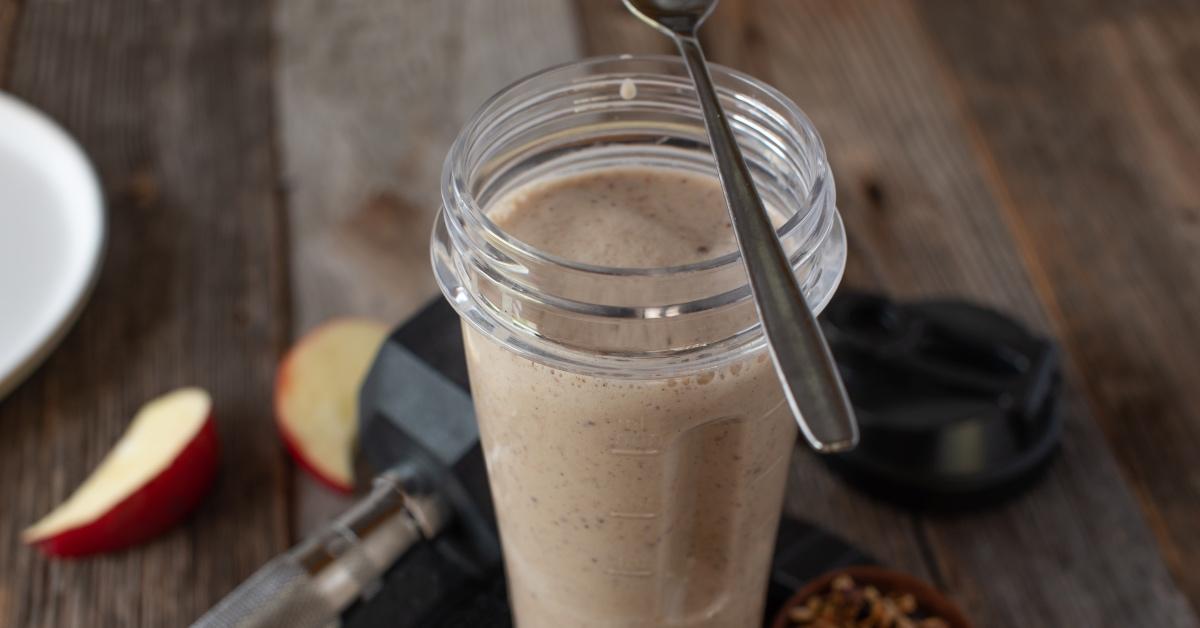 A protein shake with a spoon surrounded by apples and dumbbells. 