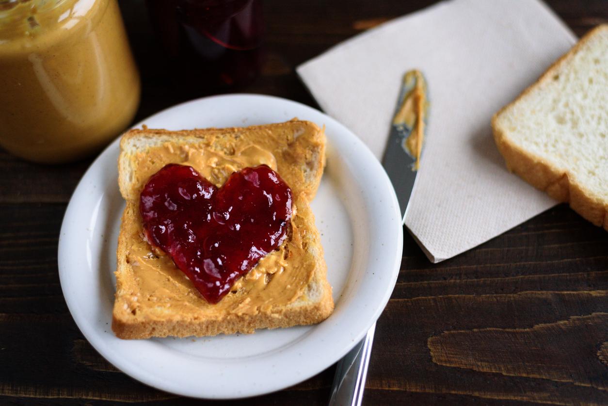 A peanut butter sandwich is pictured on a white plate with a heart-shaped dollop of jelly on top.