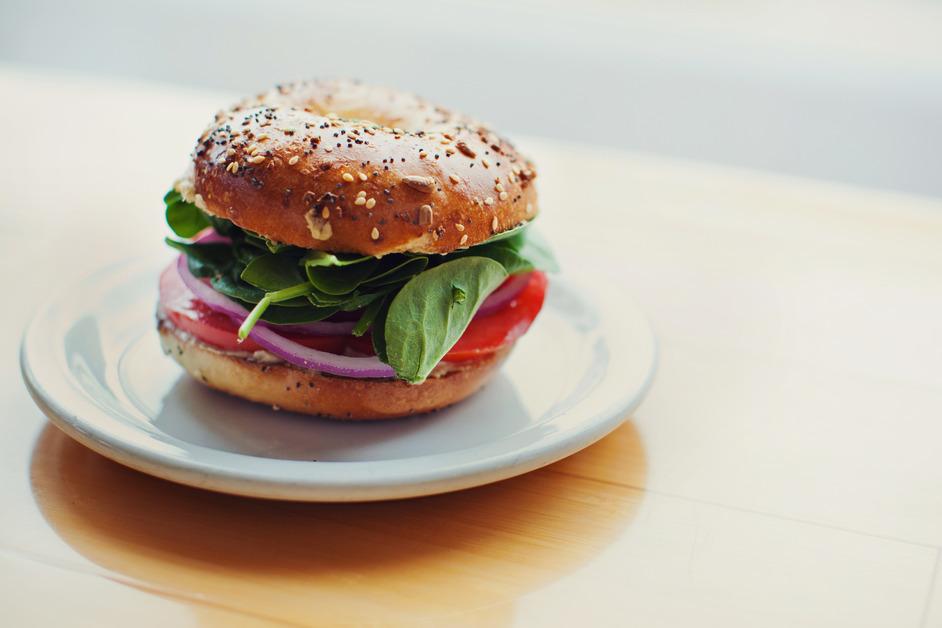 An everything bagel sandwich full of spinach, tomato, and onion sits on a plate. 