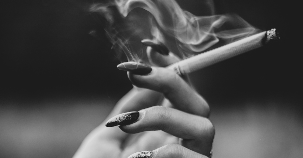 A woman with long nails holds a lit cigarette that is producing a thick smoke plume 