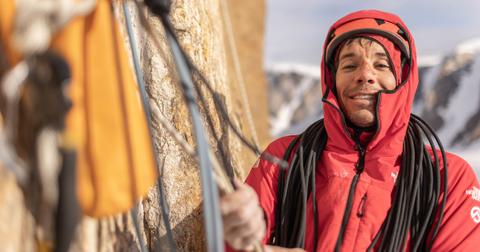 Alex Honnold has a bloody nose, and wears an orange helmet and red jacket, holding ropes on mountain, smiling at the camera.