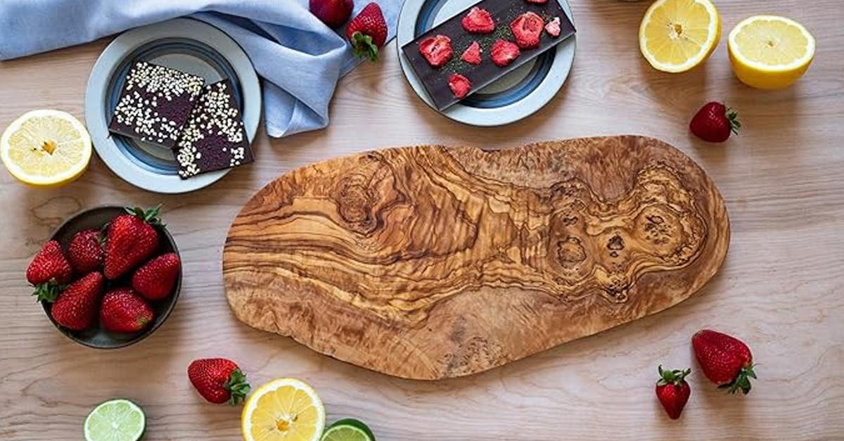 olive wood cutting board surrounded by strawberries, lemons, limes, and chocolate on a wooden table.