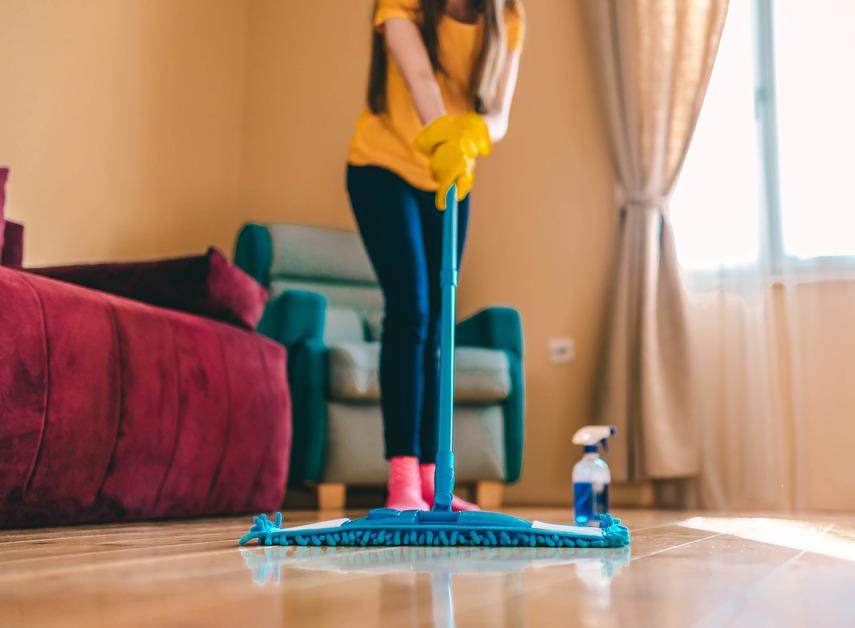 A person uses a blue mop on hardwood floors in front of a red couch and blue chair. 