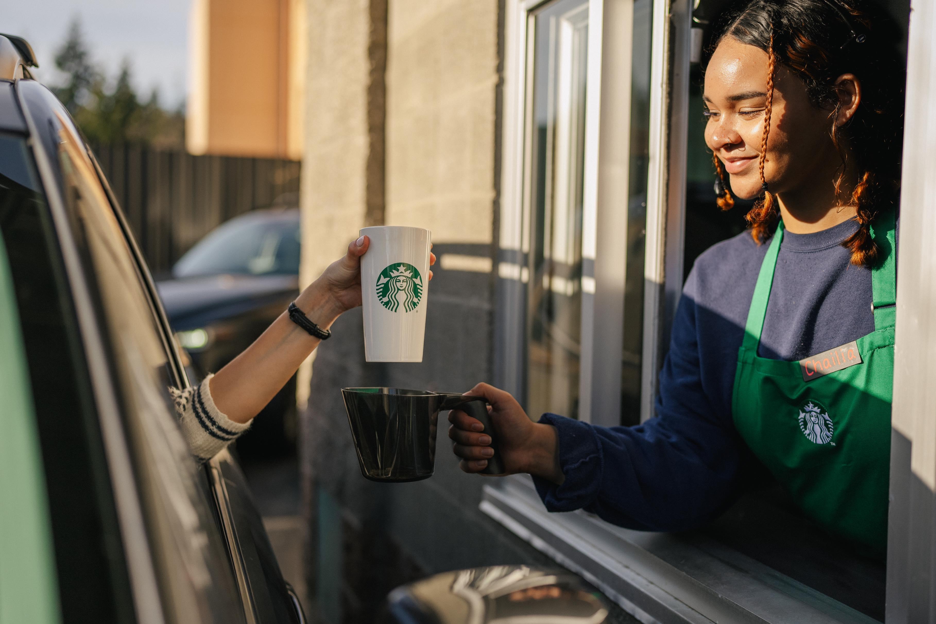 A Starbucks employee wearing a Starbucks uniform at a drive-thru hands a customer their personal white cup within a larger black container in a Starbucks store.