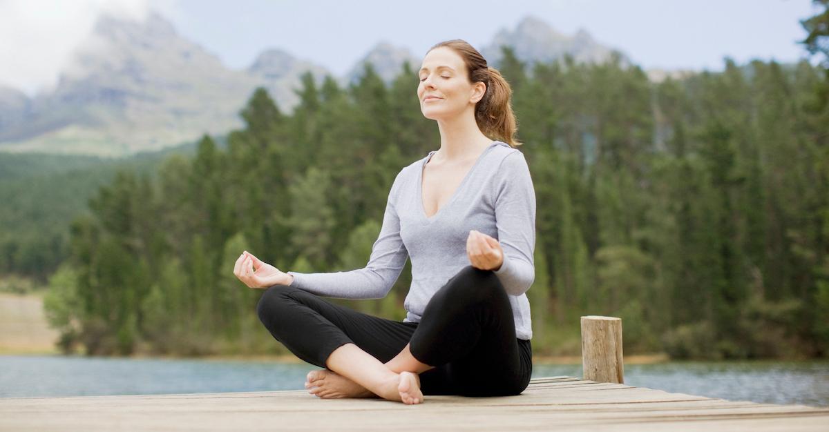 Woman sits cross-legged and meditates on deck by lake