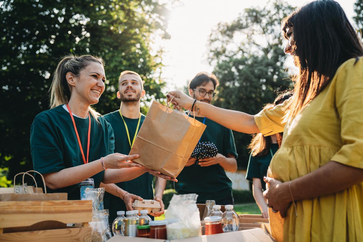 A team in green shirts hosts a community food and clothing drive, and a woman hands a brown bag to a pregnant attendee.