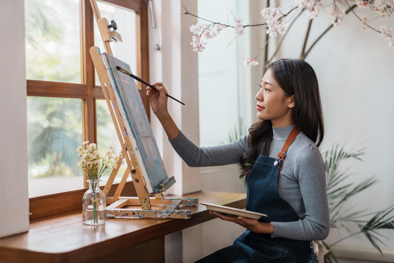 A woman wears a long sweater and apron while painting with acrylic paint.