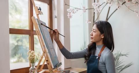 A woman wears a long sweater and apron while painting with acrylic paint.