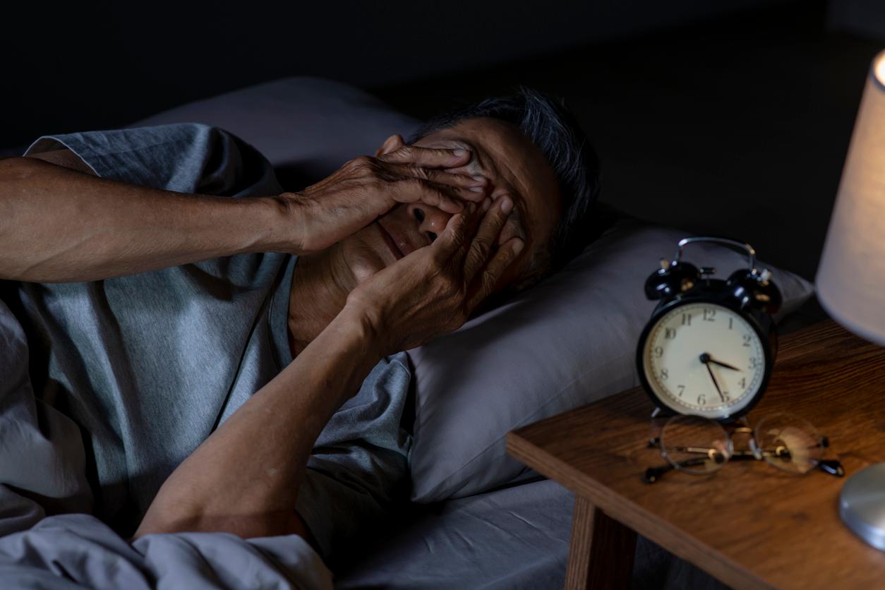 An older adult man rubs his eyes in pain in bed beside an alarm clock and glasses on the night stand.