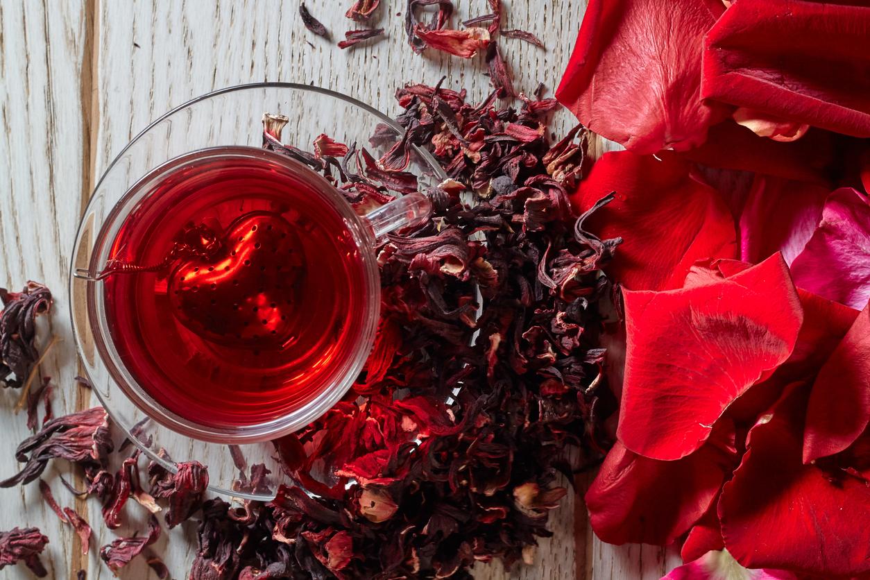 Red hibiscus tea is prepared in a glass mug on a wooden table beside rose petals.