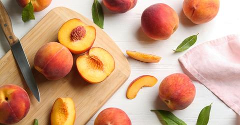 A group of peaches are cut open on a cutting board, laying beside whole peaches and green leaves on a white table cloth.