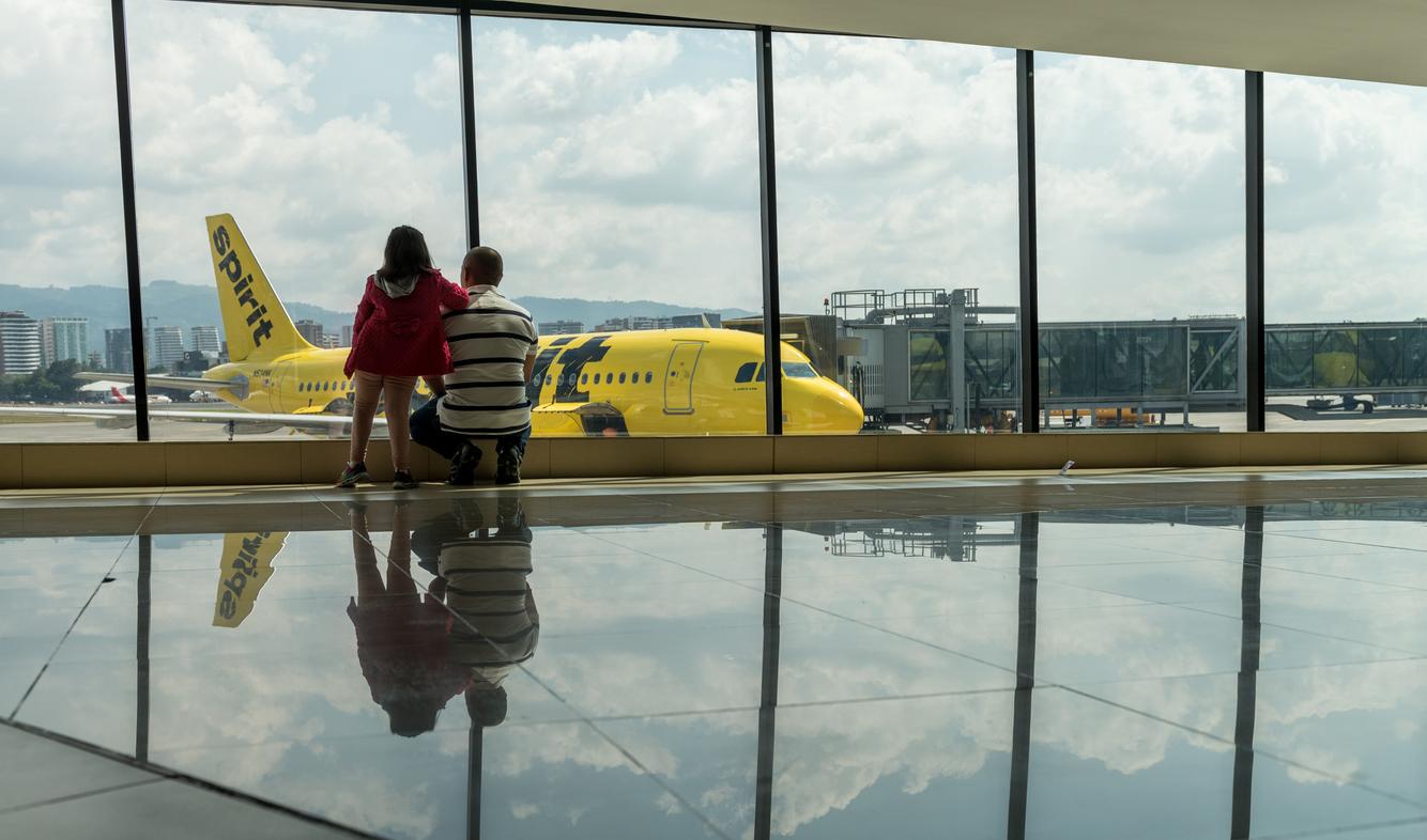 A father and daughter watch out the window of the Guatemala International Airport for their incoming Spirit Airlines airplane.