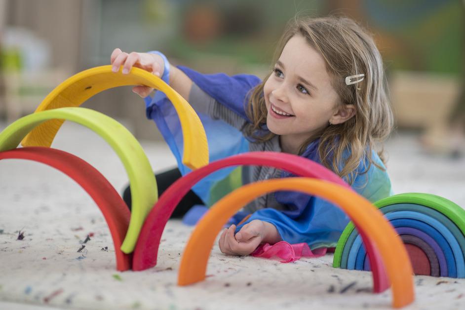 A little girl lies in the sand while stacking colorful arches. 